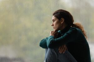 woman sits and leans her head on her arms while looking out a window wondering the differences between opiate vs opioid